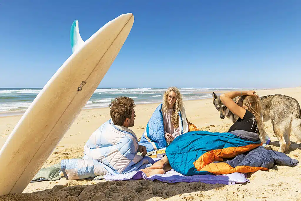 Mehrere Personen sitzen am Sandstrand eingewickelt in bunte Decken