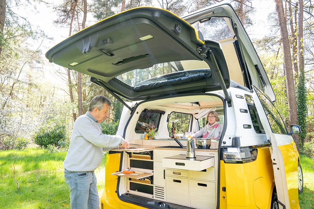 Ein Mann kocht an der Heckküche unter der Heckklappe eines leuchtend gelben Campingbusses