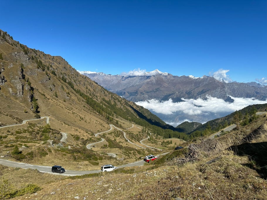 Drei Fahrzeuge auf einer Gebirgsstraße, im Hintergrund hohe, felsige Berge und Wolken