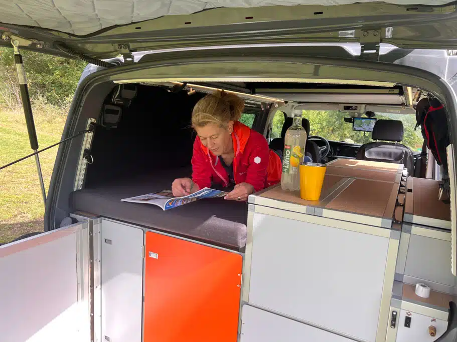 Eine Frau liegt auf einem Einzelbett im Camingbus bei geöffneter Heckklappe