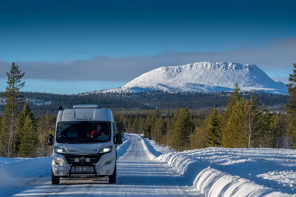 Weißer Campervan fährt auf einer Straße durch schneebedeckte Winterlandschaft, am Horizont ein schneebedeckter Berg