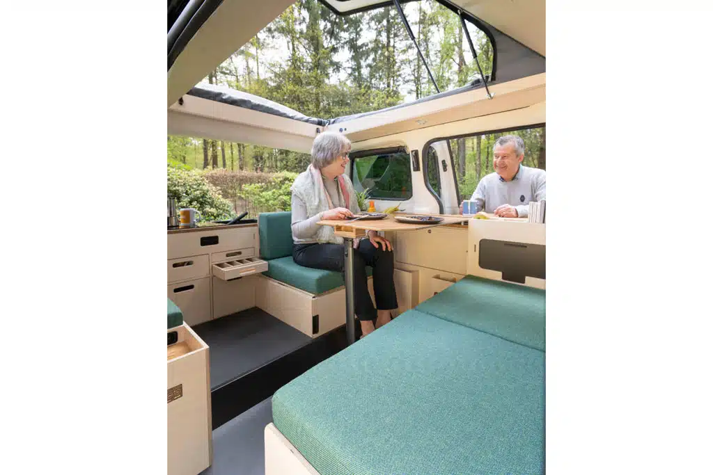 Eine Frau sitzt auf einer hölzernen Sitzgelegenheit in einem Campingbus und unterhält sich mit einem Mann, der draußen steht.