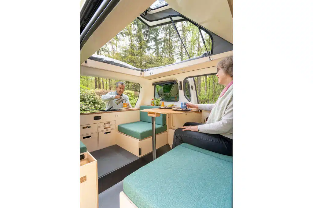 Eine Frau sitzt auf einer Sitzbank in einem Campingbus, an der Heckküche außen vor dem Fahrzeug steht ein Mann und kocht.