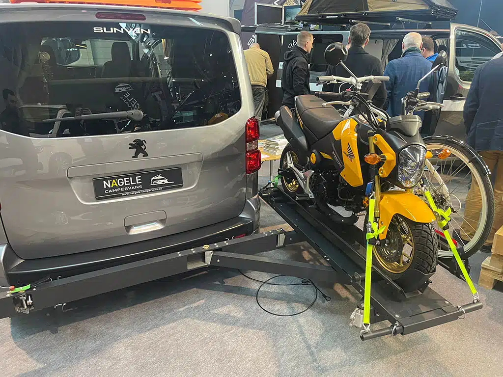 Silberner Hochdachkombi mit zur Seite weggeschwenktem Heckträger mit einem gelben Motorrad in einer Messehalle