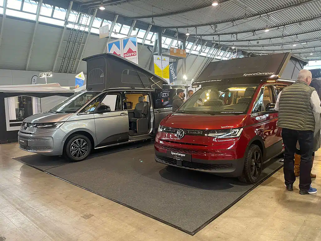 Ein roter und ein silberner Campingbus mit Aufstelldach in einer Messehalle