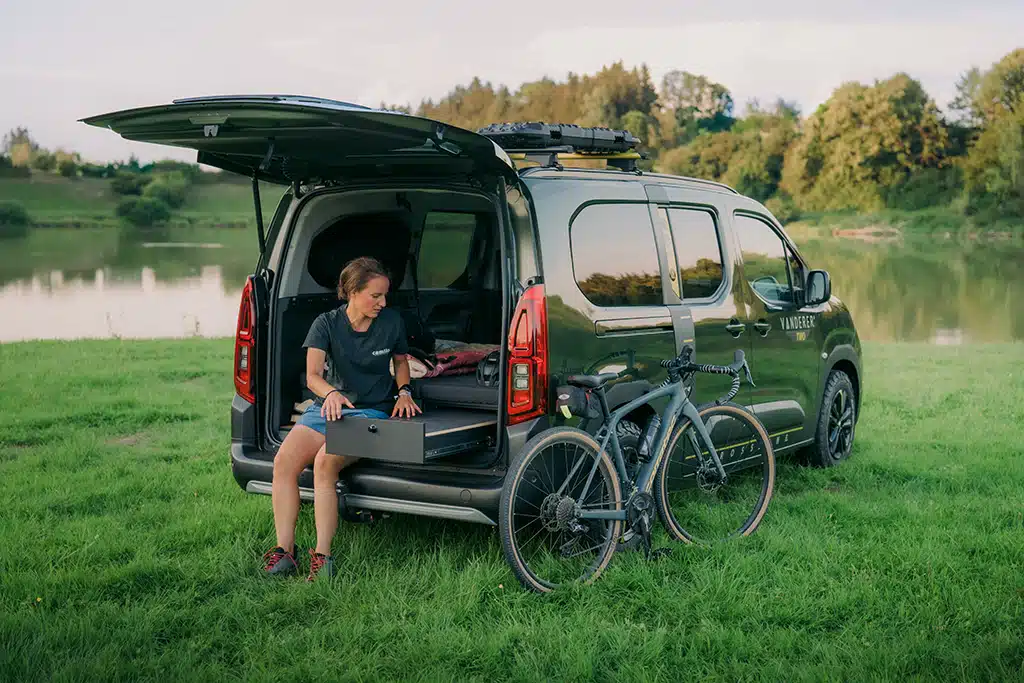 Junge Frau öffnet Schublade eines Minicampers mit Fahrrad auf einer Wiese.
