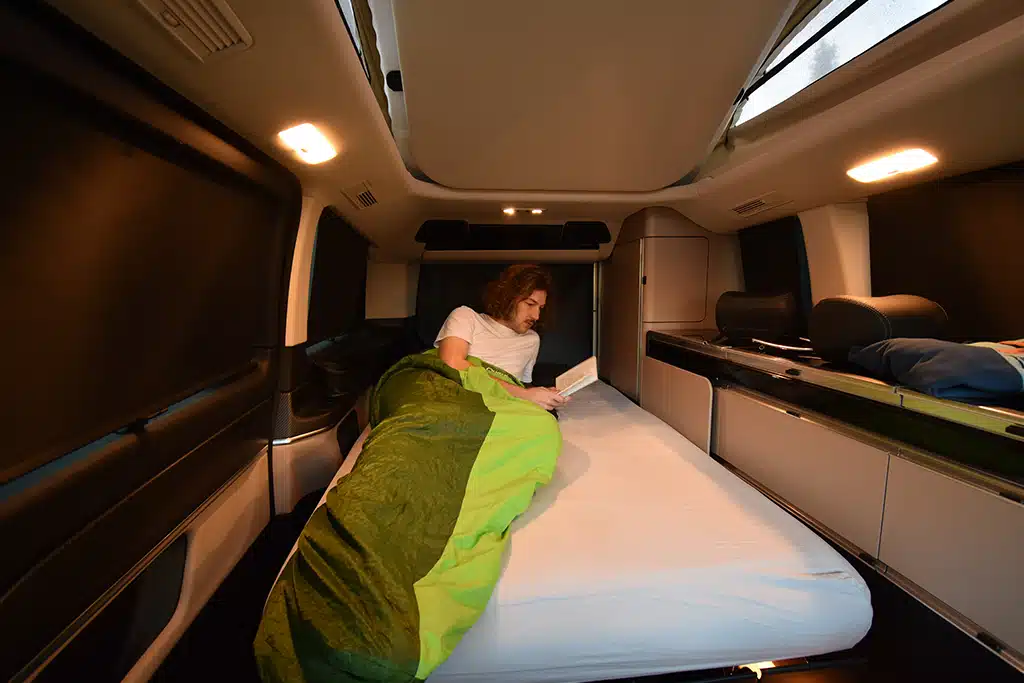 Mann mit Schlafsack und Buch auf der ausgeklappten Sitzbank des Mercedes-Benz Marco Polo.