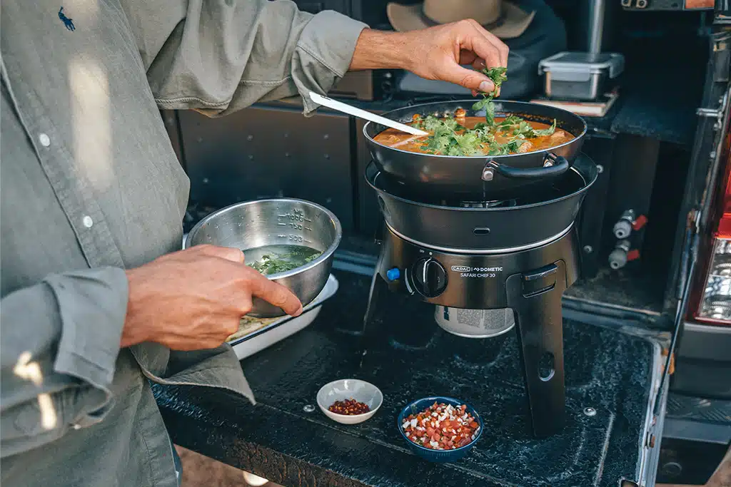Blick auf den großen Campingkocher Cadac Safari Chef auf der geöffneten Heckklappe eines Pickups, in dem eine Person, links im Bild und stark angeschnitten, eine Art Suppe oder Curry kocht. In der rechten Hand hält sie eine kleine Schüssel mit grünen Gewürzen, mit der linken Hand streut sie diese Gewürze ins Essen.