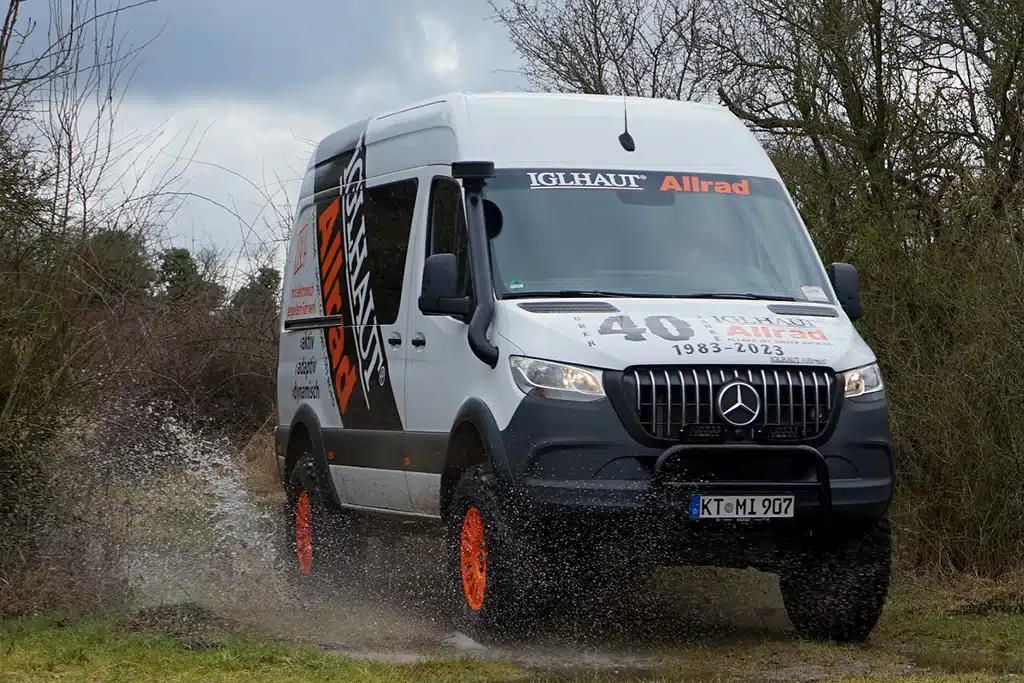 Ein Offroad-Sprinter mit martialisch anmutendem Kühlergrill und Iglhaut-Schriftzug fährt durch eine sehr nasse Wiese.
