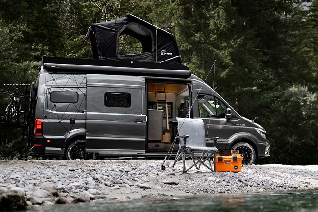 Ein MAN-Kastenwagen, ausgebaut von Loef steht mit offener Schiebetür am Ufer eines Sees. Am Fahrzeug befinden sich ein Fahrradträger mit Rädern sowie ein Dachzelt, vor dem Fahrzeug steht ein Campingstuhl, an dem eine Angel lehnt, daneben eine orangefarbene Kühlbox.