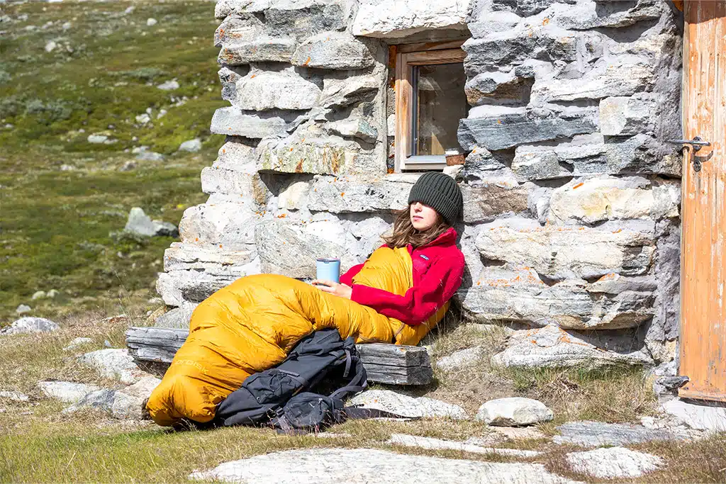 Eine junge Frau lehnt an einem Steinhaus und sitzt dabei in einem gelben Schlafsack aus der neuen Nordisk-Kollektion. In der Hand hält sie eine Tasse, ihre schwarze Mütze hat sie tief ins Gesicht gezogen.