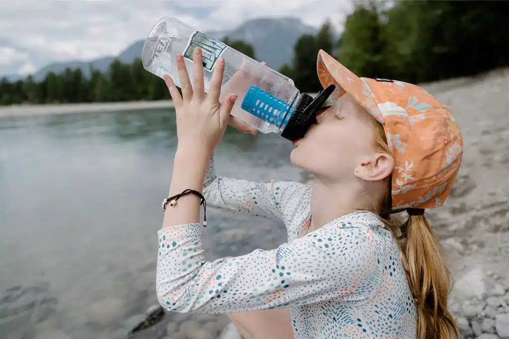 Ein Mädchen trinkt am Ufer eines Sees oder Flusses aus einer Trinkflasche, auf der der Platypus Daycap Filter installiert ist.