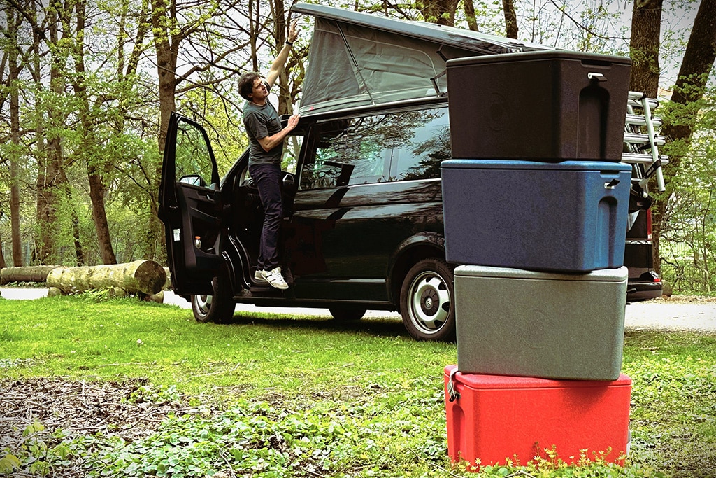 Vier Aerostuffyboxen von cagoon stehen übereinander gestapelt auf einer Wiese, im Hintergrund ein Campingbus mit aufgestelltem Hochdach.