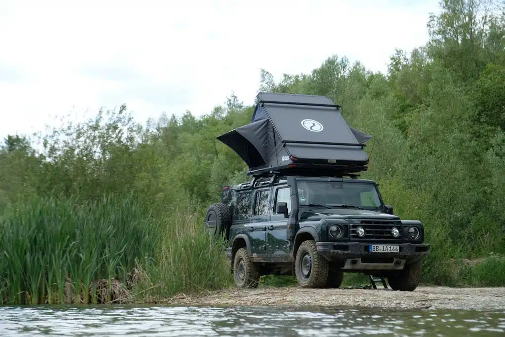 Ein Ineos Grenadier mit geöffnetem Dachzelt Alulight XL von Naturbummler steht am Ufer eines Sees.
