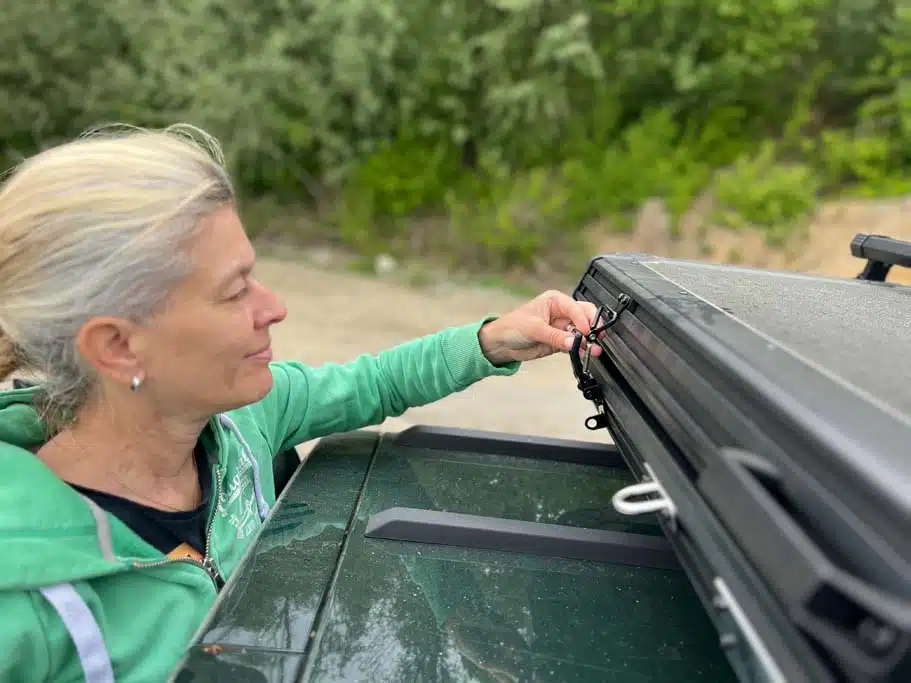 Eine Frau öffnet einen der vier Riegel des Naturbummler Dachzelts Alulight XXL, um es zu öffnen.