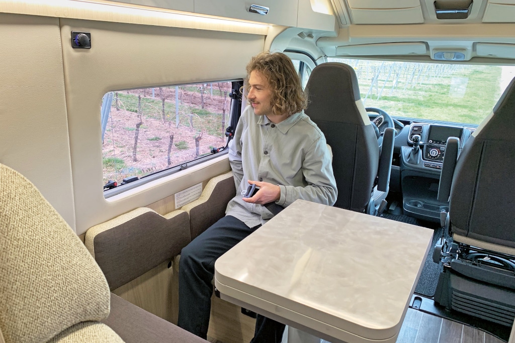 Mann sitzt auf rückwärtig augerichteten Sitz hinter Fahrersitz an zugeklapptem Esstisch