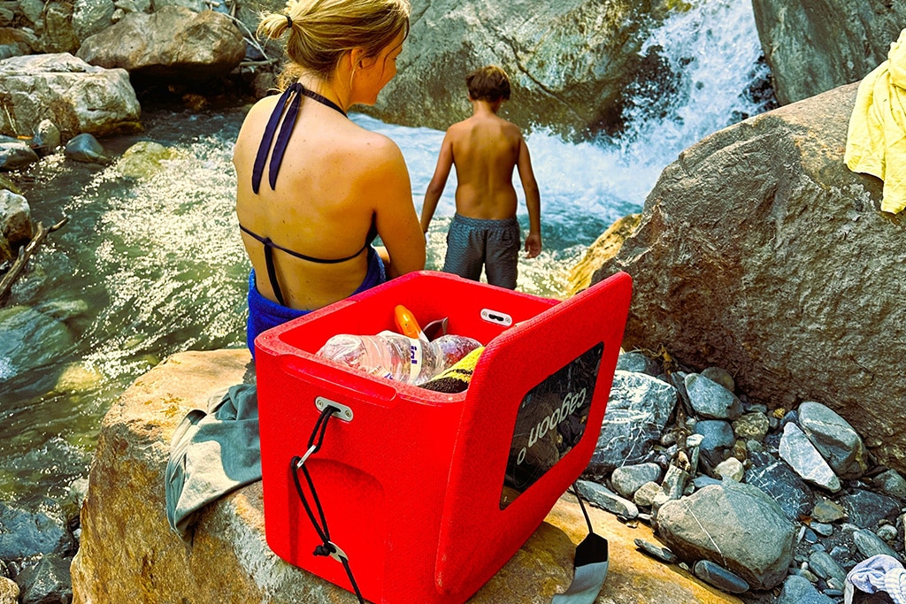 Eine Frau und ein Mann sitzen am Rand eines Wasserfalls, neben ihnen auf einem Felsen die Cagoon Passivkühlbox in Rot.