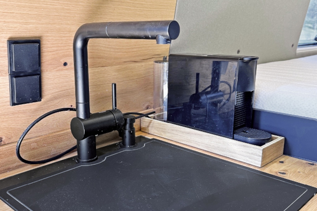 Spülabdeckung und Armatur in schwarz, daneben Kaffeemaschine ebenfalls schwarz. Spritzwand aus hellem Holz