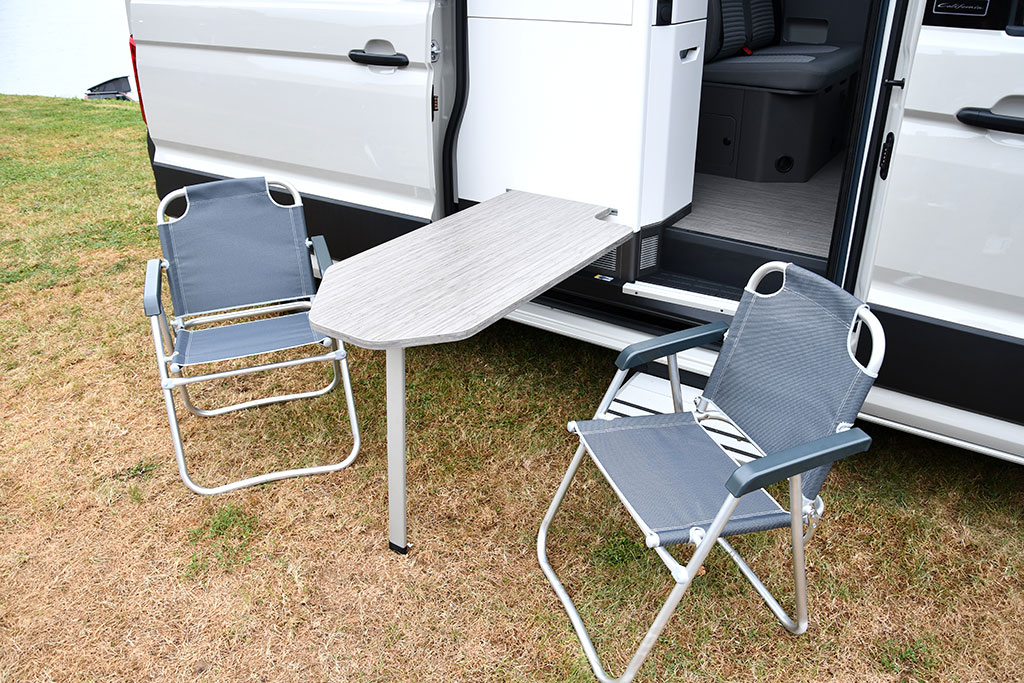 Tisch außen am Küchenblock des Campervans eingehängt, daneben zwei Campingstühle