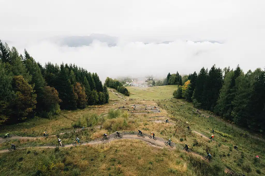 Mountainbiker fahren hintereinander auf einem schmalen Weg durch eine hügelige, waldige Herbstlandschaft.