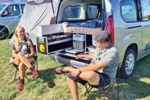 Test Ququq Campingbox: Eine junge Frau und ein junger Mann in sommerlicher Kleidung sitzen auf Campingstühlen vor der geöffneten Heckklappe eines silbernen Hochdachkombis, in dem eine Campingbox mit vorgezogenem Koch-Auszug steht.