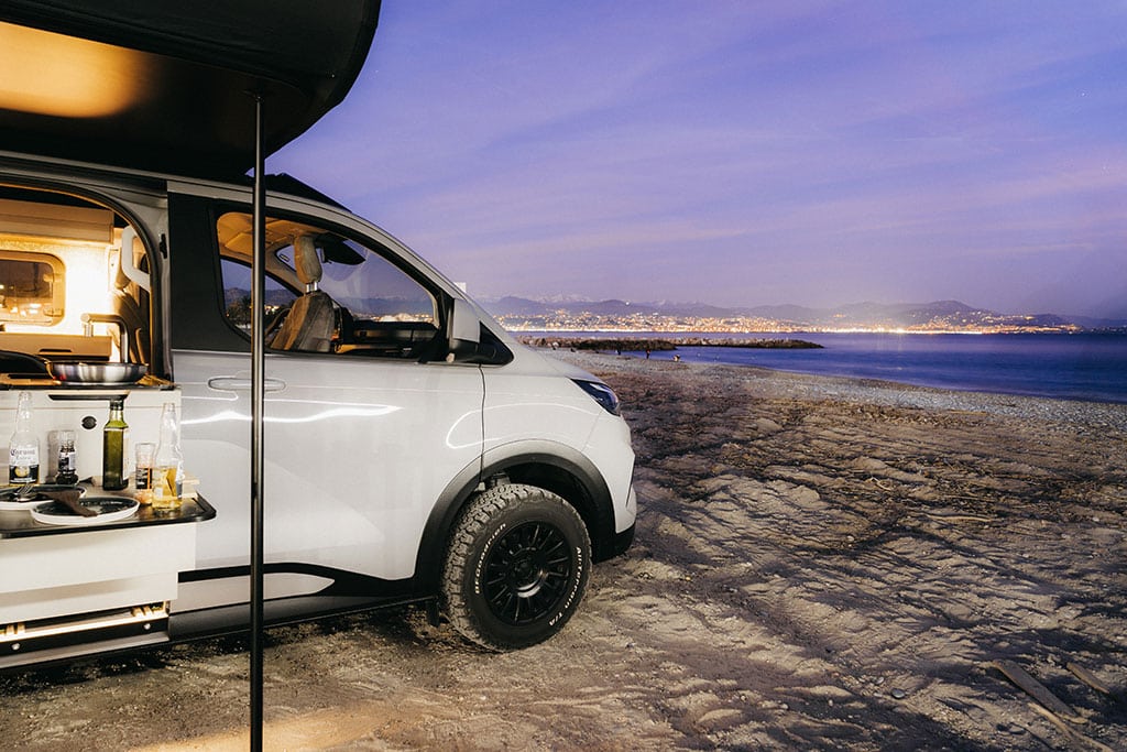 Vorderer Teil eines weißen Campingbusses auf Sand an einem Gewässer