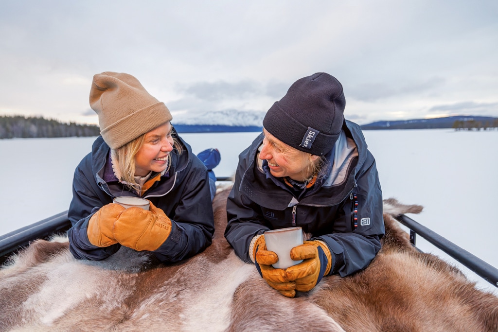 Paar liegt im Winter mit Kaffeetasse in der Hand auf Rentierfell auf dem Dach des Campers Dovra