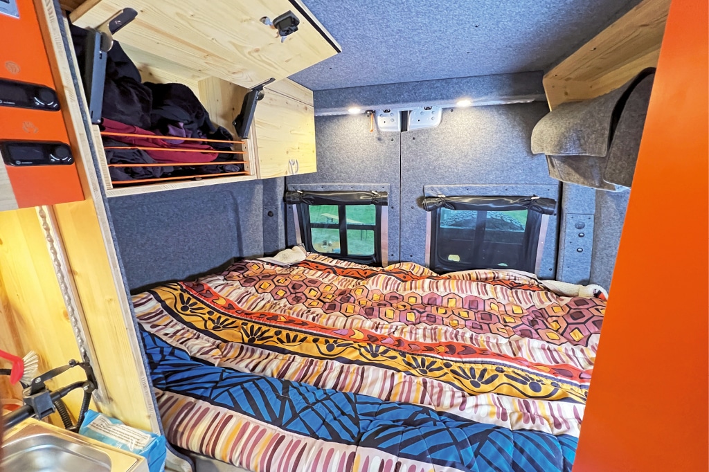 Das Querbett im Flowcamper mit poppig bunter Bettwäsche. Ein Oberschrank links oben ist offen